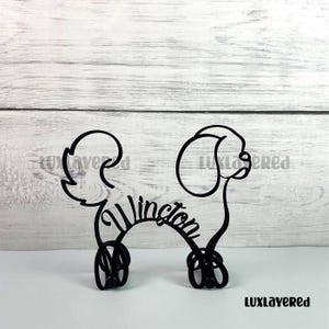 May include: Black metal dog silhouette with the name "Winston" in cursive script. The dog is depicted in a stylized, minimalist design, with a curly tail and floppy ear. The product is a decorative item.