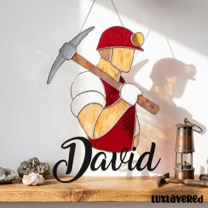 Könnte beinhalten: Glasmalerei-Kunstwerk, das einen Bergmann mit einer Spitzhacke zeigt, der einen roten Helm und eine Weste trägt. Der Name "David" ist darunter in Kursivschrift geschrieben. Eine kleine Lampe und andere Werkzeuge liegen auf einem Holzregal.