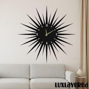 Puede incluir: Un reloj de pared negro con un diseño de rayos de sol. La esfera del reloj es negra con marcadores de hora blancos y manecillas verde neón. El reloj está montado en una pared blanca.