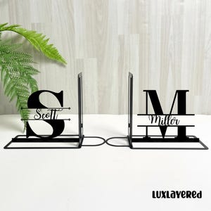 Puede incluir: Dos sujetalibros de metal negro. Cada uno presenta una letra grande con un nombre debajo. Uno tiene la letra "S" con "Scott", el otro "M" con "Miller". Los sujetalibros están sobre una superficie blanca.