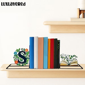 Puede incluir: Una colección de libros coloridos exhibidos en una estantería de madera, flanqueados por sujetalibros decorativos. Un sujetalibros presenta una corona floral con la letra "S", y el otro representa un libro abierto con vides frondosas. La marca "LUXLAYERED" es visible.