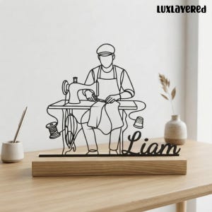 May include: A black wire-frame sculpture depicting a person sewing at a machine, mounted on a rectangular wooden base. The design includes a spool of thread, a thimble, and the name Liam in script lettering. The text Luxlayered is visible.