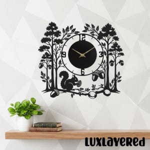 May include: Black metal wall clock featuring a forest scene with trees, acorns, and a squirrel. The clock face is black with gold hands and numerals. The clock is mounted on a white wall above a wooden shelf.