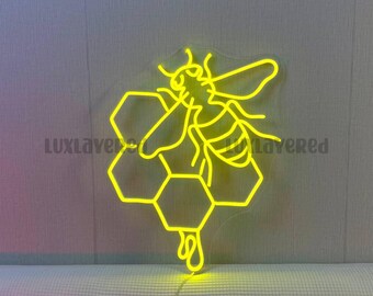 May include: A bright yellow neon sign featuring a stylized bee perched on a cluster of hexagonal honeycomb cells. The design is mounted on a clear acrylic backing, with the text LUXLAYERED visible in the center and bottom right corner.