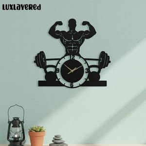 May include: Black wall clock featuring a muscular bodybuilder silhouette with raised arms. The clock face is round with gold hands and numbers. Dumbbells and kettlebells flank the clock, creating a fitness-themed design. The text "LUXLAYERED" is at the top.