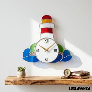 Könnte beinhalten: Eine Leuchtturm-Wanduhr mit weißem Zifferblatt und goldenen Zeigern. Der Leuchtturm hat rote, weiße und gelbe Glasmalereien. Die Uhr ist an einer weißen Wand über einem Holzregal befestigt. Das Design der Uhr umfasst blaue und grüne Wellen.