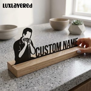 Könnte beinhalten: Eine schwarze Silhouette einer Person, die Mundharmonika spielt, befestigt auf einem hellen Holzsockel. Die Silhouette zeigt den Text CUSTOM NAME in fetten Großbuchstaben. Das Objekt steht auf einer grauen, gesprenkelten Oberfläche.