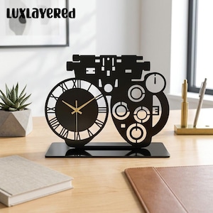 May include: A black metal desk clock shaped like an optometrist's phoropter. The clock face features Roman numerals and gold-colored hands. The design includes circular cutouts and mechanical details, all mounted on a rectangular black base.