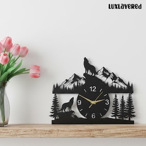 May include: Black wall clock with a mountain and wolf design. The clock has a round face with gold hands and numbers. The design includes silhouettes of mountains, trees, and wolves. The text "LUXLAYERED" is at the top.