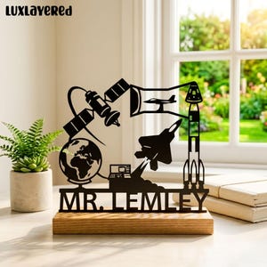 May include: Black metal desk decor featuring a space theme with a globe, satellite, rocket, and jet. The base is made of wood, and the text "MR. LEMLEY" is displayed. A small plant in a pot is on the left.