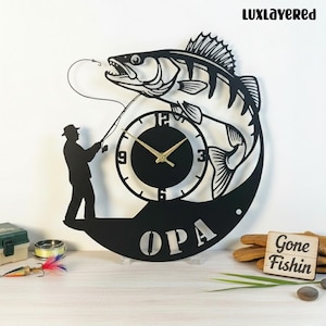 May include: Black metal wall clock featuring a fish design with a fisherman silhouette. The clock face is integrated into the fish's body, with the word "OPA" at the bottom. The clock has gold-colored hands.