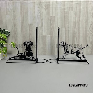 Puede incluir: Juego de sujetalibros de metal negro con diseño de perros. Uno muestra un perro tumbado y el otro un perro en movimiento. Los sujetalibros tienen una base rectangular y un soporte vertical, unidos por un diseño circular.