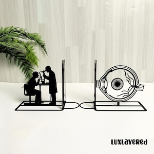May include: Black metal bookends featuring an ophthalmologist examining a patient and a detailed eye anatomy illustration. The bookends are connected by a curved bar. The word "LUXLAYERED" is printed in black.