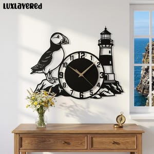 May include: A black and white wall clock featuring a puffin standing on a rock next to a lighthouse. The clock face is circular with black numbers on a white border and a solid black center. The text LUXLAYERED is visible in the top left corner.