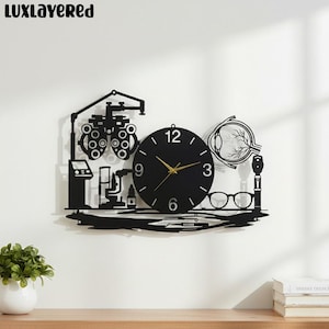 Puede incluir: Reloj de pared negro con manecillas doradas, con un tema de oftalmología. El diseño del reloj incluye una tabla optométrica, gafas y una máquina de examen ocular. El reloj está montado en una pared blanca.