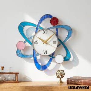 May include: A decorative wall clock designed as an atom, with a white clock face and gold hands. The atom's structure is made of blue and red stained glass. The clock adds a unique and stylish element to any interior space.