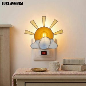 May include: A decorative night light shaped like a sun and cloud. The sun has yellow rays and a yellow center, with a light bulb inside. The cloud is white and blue. The night light is plugged into a wall outlet and has a red on/off switch. The brand name "LUXLAYERED" is at the top.