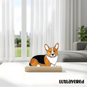 May include: A stained glass corgi dog figurine in shades of orange, black, and white, resting on a wooden base. The dog is in a lying down position. The figurine is displayed on a white table in front of a window.