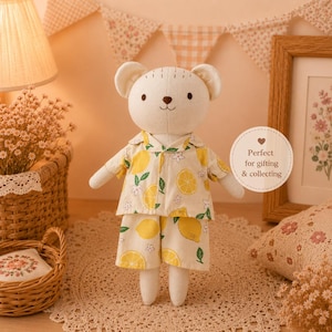 Puede incluir: Un muñeco de oso blanco de peluche que lleva un pijama amarillo con estampado de limones. El muñeco tiene una cara cosida y orejas pequeñas. Un letrero redondo con el texto "Perfect for gifting & collecting" es visible.