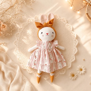 May include: A plush rabbit doll with white fur, wearing a mustard yellow headband with a bow, and a white dress featuring vertical red stripes and small floral patterns. The doll has embroidered facial features and mustard yellow feet.