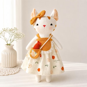 May include: A plush cat doll with white fur, orange patches, and a matching orange bow on its head. It wears a cream-coloured dress with an apple print, a mustard yellow apron with white lace trim, and a small crossbody bag containing a red fish toy.