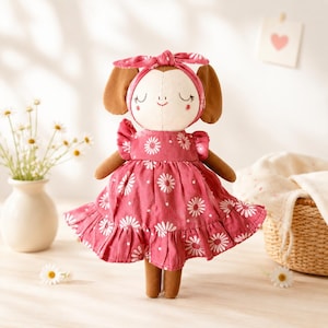 Puede incluir: Una muñeca de peluche con extremidades marrones y cara color crema con ojos cerrados y boca pequeña bordados. La muñeca viste un vestido rosa oscuro con estampado de margaritas blancas, mangas con volantes, dobladillo con volantes y diadema a juego.