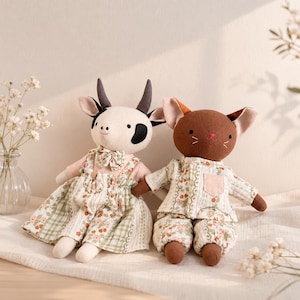 Puede incluir: Dos muñecas de tela hechas a mano: una vaca y un ratón. La muñeca de vaca tiene una cara blanca con manchas negras y cuernos, con un vestido rosa y floral. La muñeca de ratón es marrón con orejas grandes, con un pijama floral.