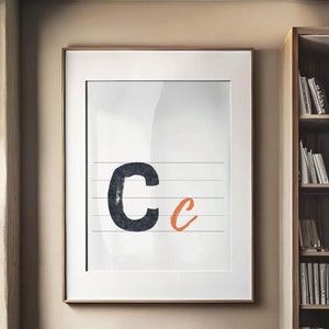 Op de afbeelding: Een ingelijst print met een zwart-witte letter "C" en een oranje letter "c" op een witte achtergrond met horizontale lijnen. De print hangt aan een beige muur naast een boekenkast en een groene fauteuil.