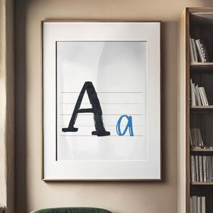 Op de afbeelding: Een ingelijst afdruk van de letter "A" in zwarte inkt op een witte achtergrond met een blauwe kleine letter "a" ernaast. De afdruk hangt aan een beige muur naast een boekenkast.