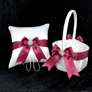 May include: A white satin ring pillow and flower girl basket set. Both items feature a wide burgundy ribbon with a sparkling rhinestone embellishment. The pillow has a loop for rings. The basket has a white handle and burgundy bows.