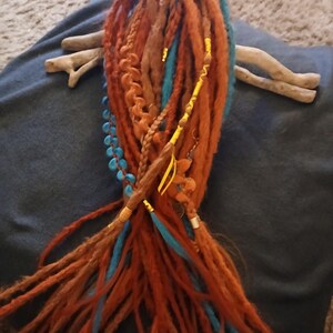 May include: Handmade dreadlocks in shades of orange, brown, and teal. The dreads are decorated with a yellow ribbon, beads, and metal accents. The dreadlocks are arranged on a dark blue surface. A pair of driftwood pieces are also present.