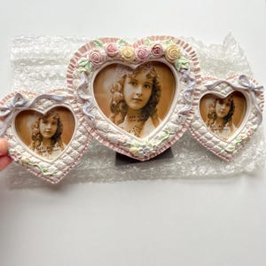 Puede incluir: Un marco de fotos triple en forma de corazón con una estética vintage. Los marcos son blancos con ribetes rosas, adornados con rosas y lazos. Cada marco contiene un retrato en sepia de una niña. El marco está sobre plástico de burbujas.