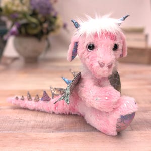 May include: A pink plush dragon toy with a fluffy mane and glitter accents. The dragon has blue horns and iridescent wings and tail details. The toy is sitting on a wooden surface.