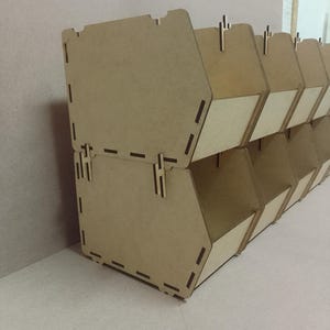 May include: A set of stacked, light brown wooden storage bins. Each bin has a trapezoidal shape with a rectangular opening and a small, light-colored interior panel. The bins are designed to interlock and are suitable for organizing small items.