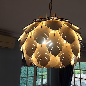 Puede incluir: Una lámpara colgante decorativa con diseño de hojas. La luz está hecha de madera de color claro y cuelga de una cadena. Las hojas están dispuestas en capas alrededor de la fuente de luz, creando un brillo cálido.