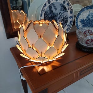 Puede incluir: Una lámpara de madera con forma de alcachofa con una luz blanca cálida. La lámpara está sobre una mesa de madera con un cable blanco.