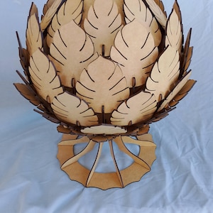 May include: A decorative wooden sculpture resembling a stylized artichoke or flower. The sculpture is made of light brown wood, with intricate leaf-like cutouts forming its outer layers. It sits on a circular base supported by curved legs, creating a unique, artistic piece.