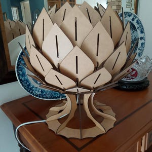 May include: A decorative table lamp with a unique design. The lamp features a layered, petal-like structure made of light brown wood. The base is also made of wood and has an intricate design. The lamp is on a wooden table.