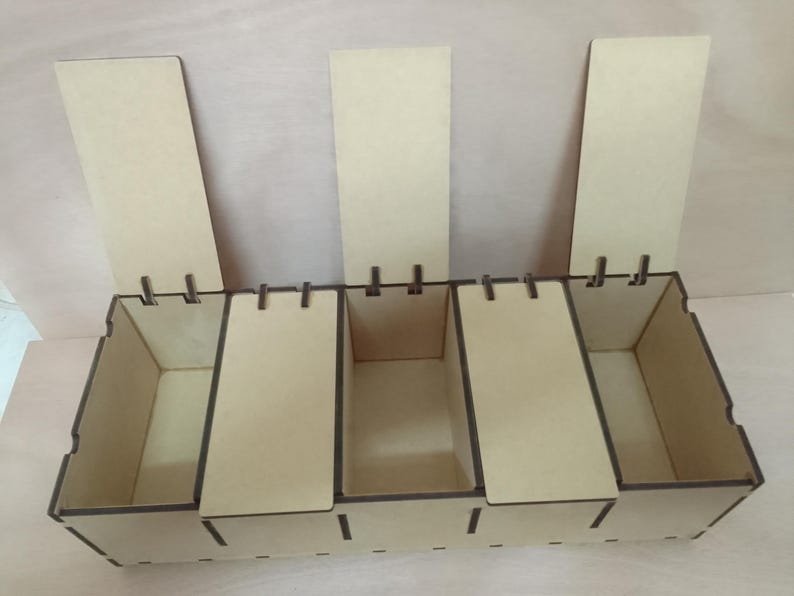 May include: A wooden storage organiser with five compartments and hinged lids. The natural wood colour is accented by darker edges. The lids are open, revealing the interior of the storage boxes. The organiser is designed for small items.