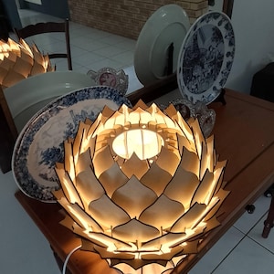 Può includere: Una lampada in legno a forma di fiore di loto che emette una luce calda. La lampada è realizzata in legno chiaro a strati e poggia su un tavolo di legno. Piatti decorativi sono visibili sullo sfondo.