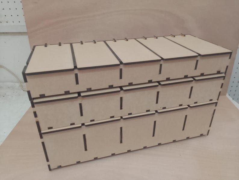 May include: A three-tiered, light brown wooden storage box with multiple compartments. Each tier has individual pull-out drawers. The box is constructed from laser-cut wood, with visible slots and a clean, geometric design. The box is suitable for organizing small items.