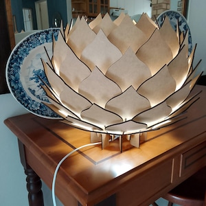 May include: A decorative wooden lamp shaped like a lotus flower, with a warm, glowing light emanating from within. The lamp is made of layered, light-colored wood with dark edges, and sits on a wooden table. A white power cord extends from the lamp.