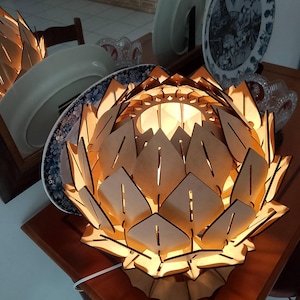 Puede incluir: Una lámpara de madera con forma de flor de loto que emite un brillo cálido. La lámpara está hecha de madera clara y tiene un diseño detallado y en capas. Está sobre una mesa de madera oscura, con platos decorativos en el fondo.