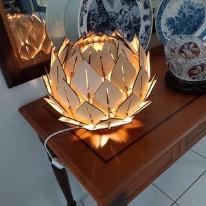 May include: A wooden lotus flower-shaped lamp with a warm glow, sitting on a brown table. The lamp's design features leaf-shaped cutouts. Decorative plates with blue and white patterns are in the background.