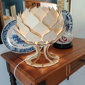 May include: A wooden lotus flower-shaped lamp with a warm glow. The lamp has a detailed design with multiple petals and a sturdy base. The lamp is sitting on a wooden table. The lamp is plugged in with a white cord.