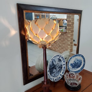 以下が含まれることがあります： 木製のテーブルランプで、白い花の形をしたシェードが付いています。シェードは、円形に配置された多くの個別の木片でできています。ランプは点灯しており、暖かな光を放っています。ランプは、後ろに鏡のある木製のテーブルの上に置かれています。