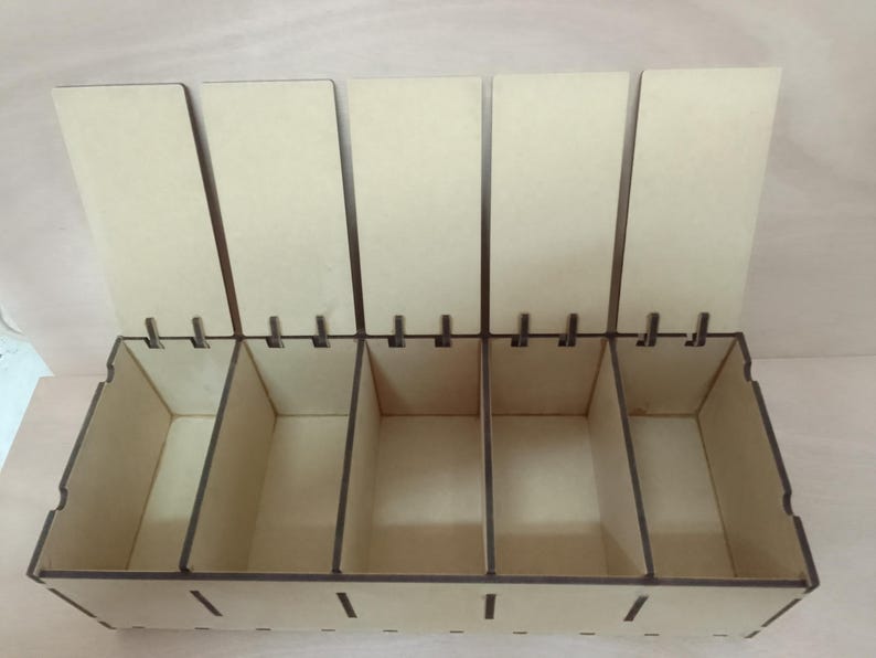 May include: A wooden storage box with five compartments and hinged lids. The box is made of light-coloured wood with dark brown edges. The lids are open, revealing the empty compartments inside. The box is designed for organising small items.