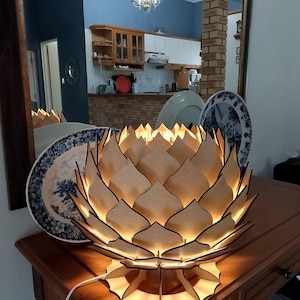 Könnte beinhalten: Eine holzfarbene Lotusblüten-Lampe mit warmem Licht. Die Lampe steht auf einem Holztisch, ein weißes Kabel führt heraus. Dekorative Teller mit blau-weißen Mustern befinden sich im Hintergrund.