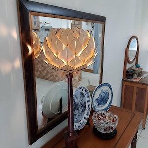 以下が含まれることがあります： 木製のテーブルランプで、ユニークな薄い茶色の花の形をしたシェードが付いています。シェードは、葉の形にカットされた多くの個別の木片でできています。ランプは点灯しており、温かい光を放っています。ランプは、装飾的なボウルと2つのプレートが置かれた木製のテーブルの上に置かれています。