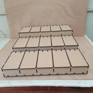 May include: Three-tiered, light brown wooden display shelves. Each shelf has multiple rectangular compartments. The shelves are arranged in a stepped configuration. The top shelf is the smallest, the bottom the largest.